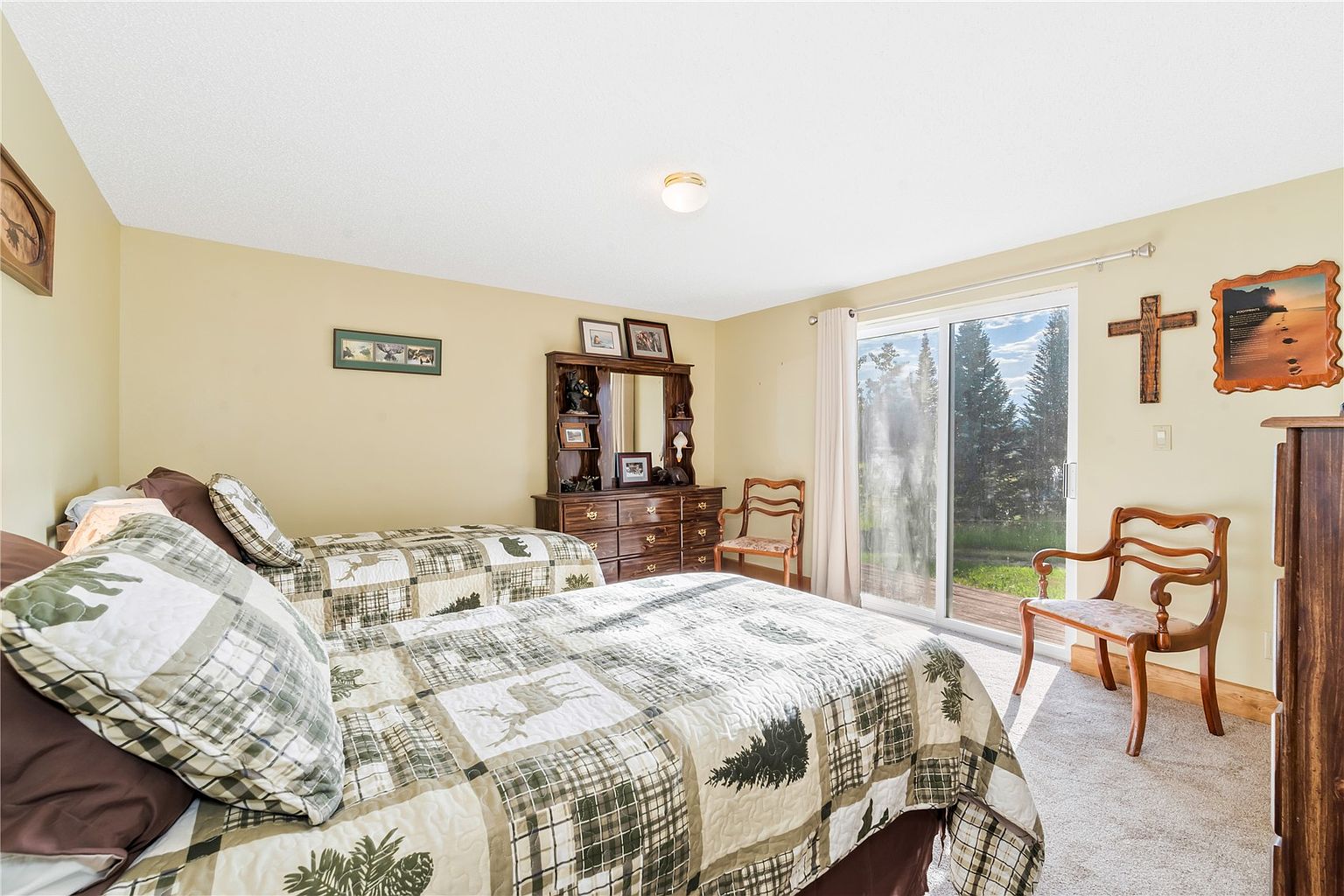 This cozy bedroom features two twin beds dressed in rustic, nature-themed patterned bedding. A wooden dresser with a mirror sits between the beds, while a wooden chair is positioned near the sliding glass doors that provide natural light and a view of the outdoors. The room is decorated with wall art, including a cross and a framed landscape, creating a warm and inviting atmosphere.