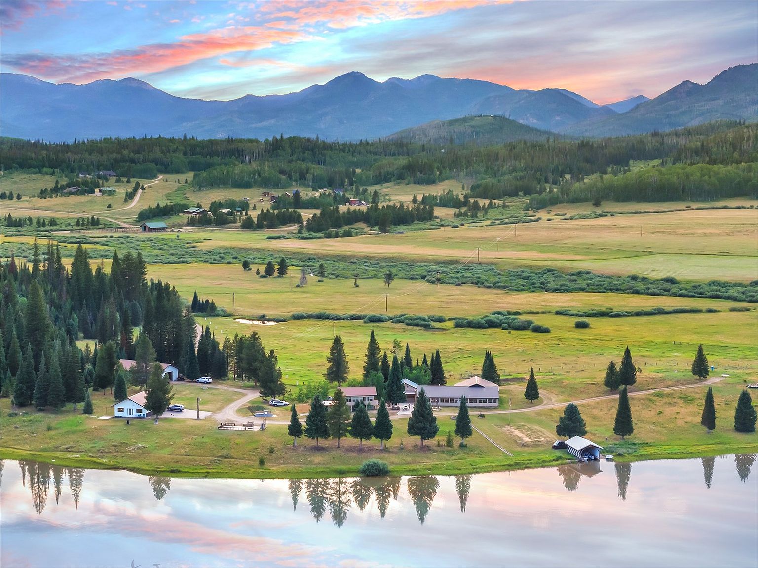 This expansive aerial view captures a serene lakeside property nestled within a vast, rolling valley surrounded by majestic mountains. The scene features a collection of buildings, including a main residence and a boat house, set against a backdrop of lush green meadows, dense pine forests, and a tranquil lake reflecting the soft colors of a sunset sky. The perspective provides a comprehensive look at the property's generous acreage and its peaceful, secluded natural setting.