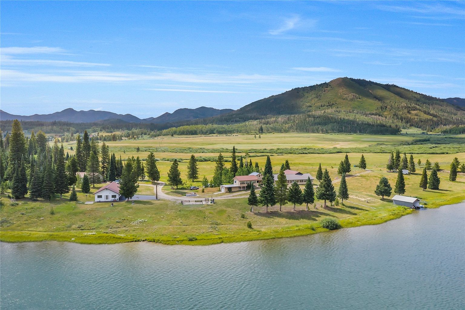 This expansive aerial view captures a serene lakeside property nestled in a vast, grassy valley surrounded by rolling hills and distant mountains. The site features multiple structures, including a primary residence and a detached garage, all set against a backdrop of mature evergreen trees and a calm, blue lake. The perspective provides a comprehensive look at the property's generous acreage, natural landscape, and peaceful, secluded atmosphere.