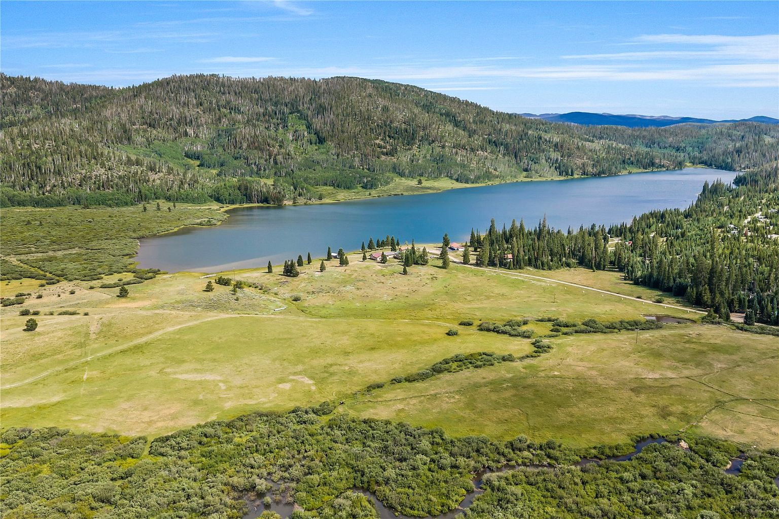 This expansive aerial view captures a serene mountain landscape featuring a large, deep blue lake nestled against a backdrop of dense, forested hills. In the foreground, a lush green meadow is bisected by a winding stream, leading toward a small cluster of buildings situated near the shoreline. The scene conveys a sense of peaceful seclusion and natural beauty, ideal for a recreational or retreat-style property.