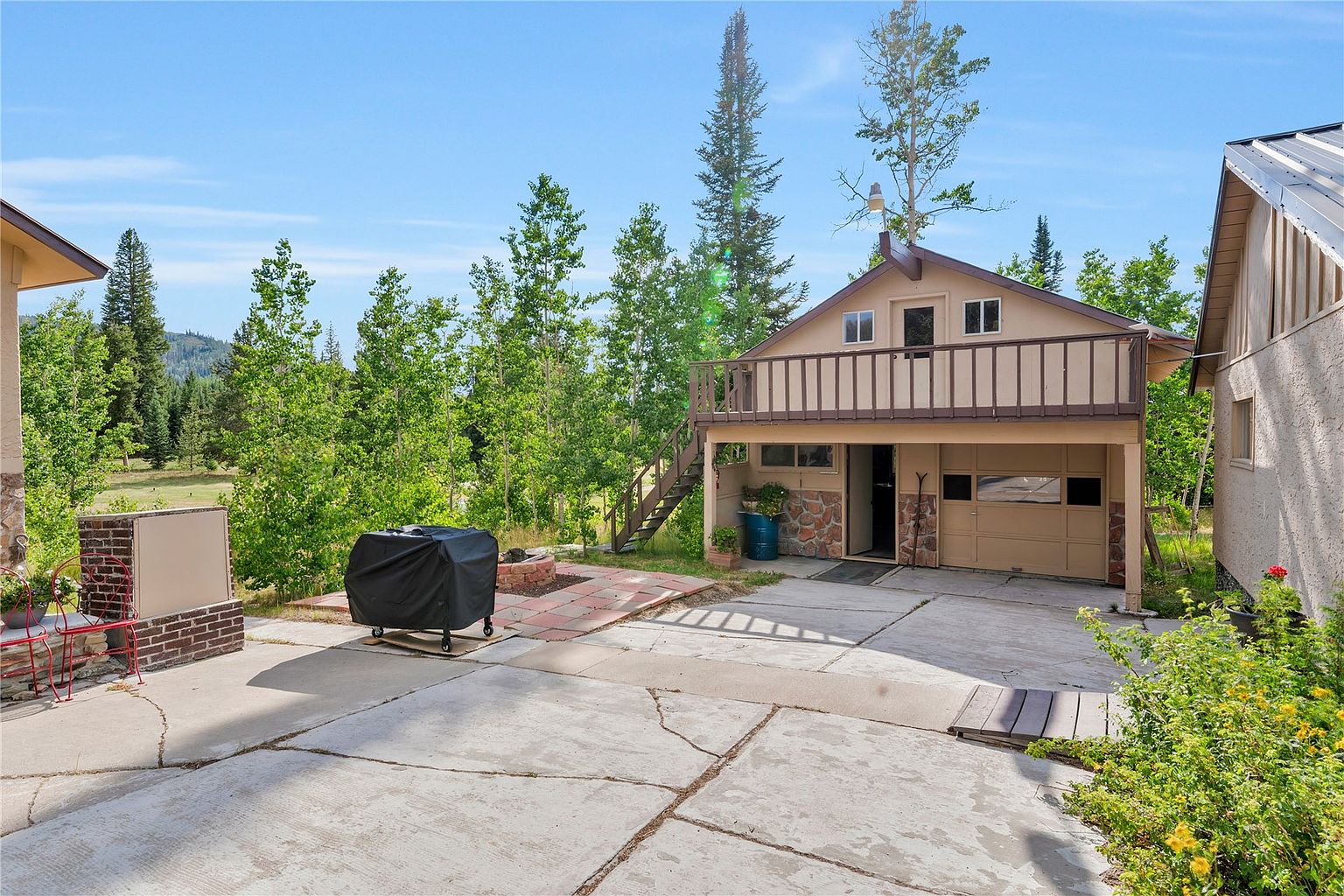 This image captures a detached two-story garage structure with a balcony on the upper level, accessed by an exterior wooden staircase. The lower level features a garage door and stone-accented walls, while the surrounding area includes a concrete driveway, a covered grill, and a small outdoor seating area. The setting is lush and wooded, providing a rustic, mountain-retreat aesthetic.