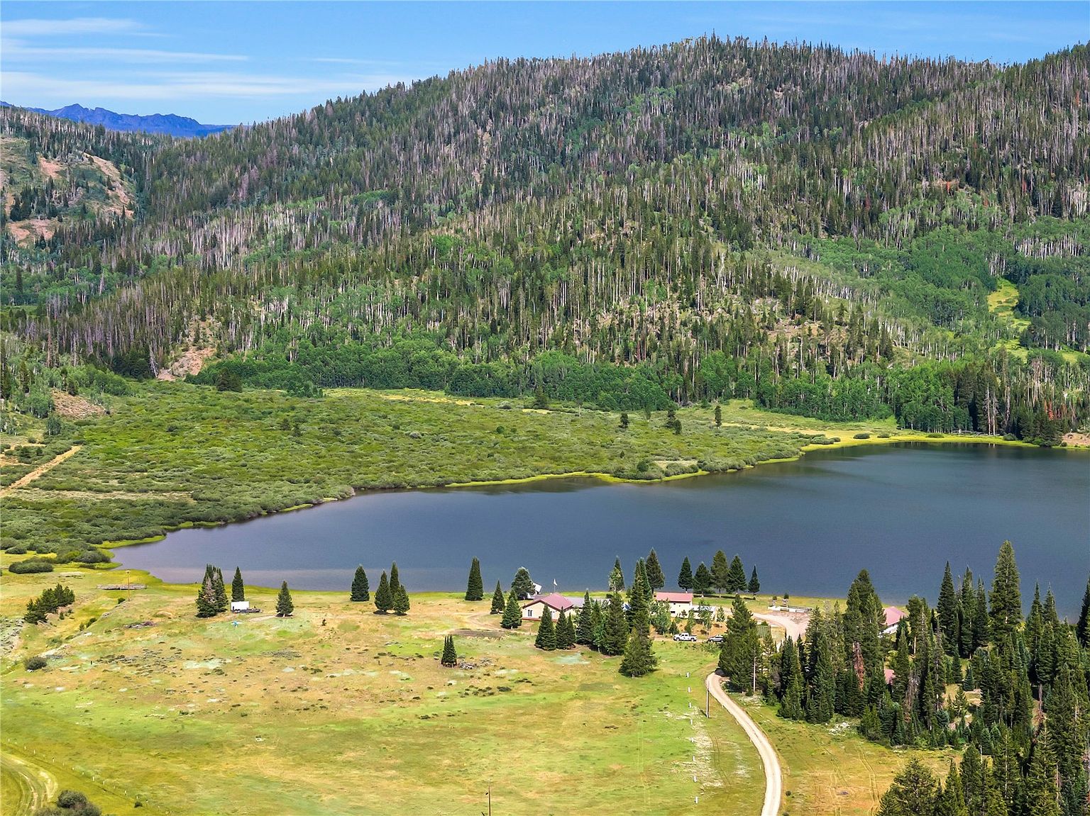 This high-angle aerial view captures a serene mountain property nestled beside a large, calm lake. The landscape features expansive grassy meadows, scattered evergreen trees, and a cluster of buildings situated near the water's edge, with a dirt road winding through the foreground. The backdrop is dominated by a vast, forested mountain slope under a clear blue sky, emphasizing the property's secluded and natural setting.