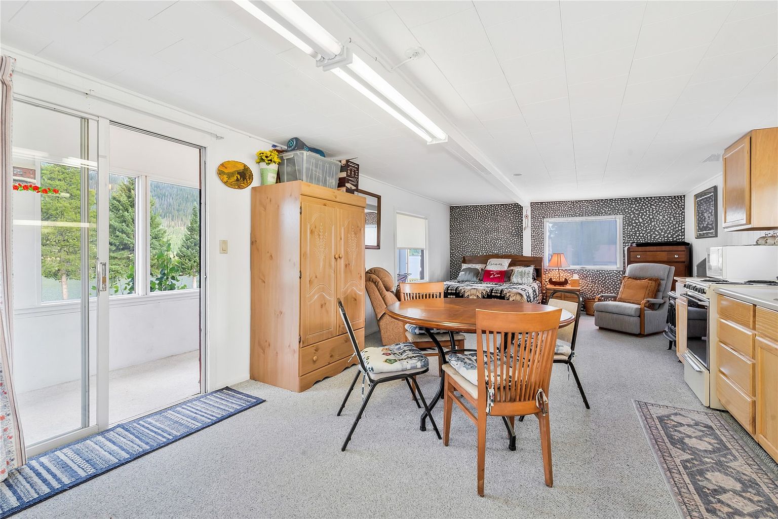 This open-concept living and dining area features a neutral color palette with a distinctive patterned accent wall behind the bed. The space includes a light wood armoire, a round dining table with wooden chairs, and a cozy seating area with a grey armchair. Large sliding glass doors provide access to an outdoor space, allowing natural light to fill the room.