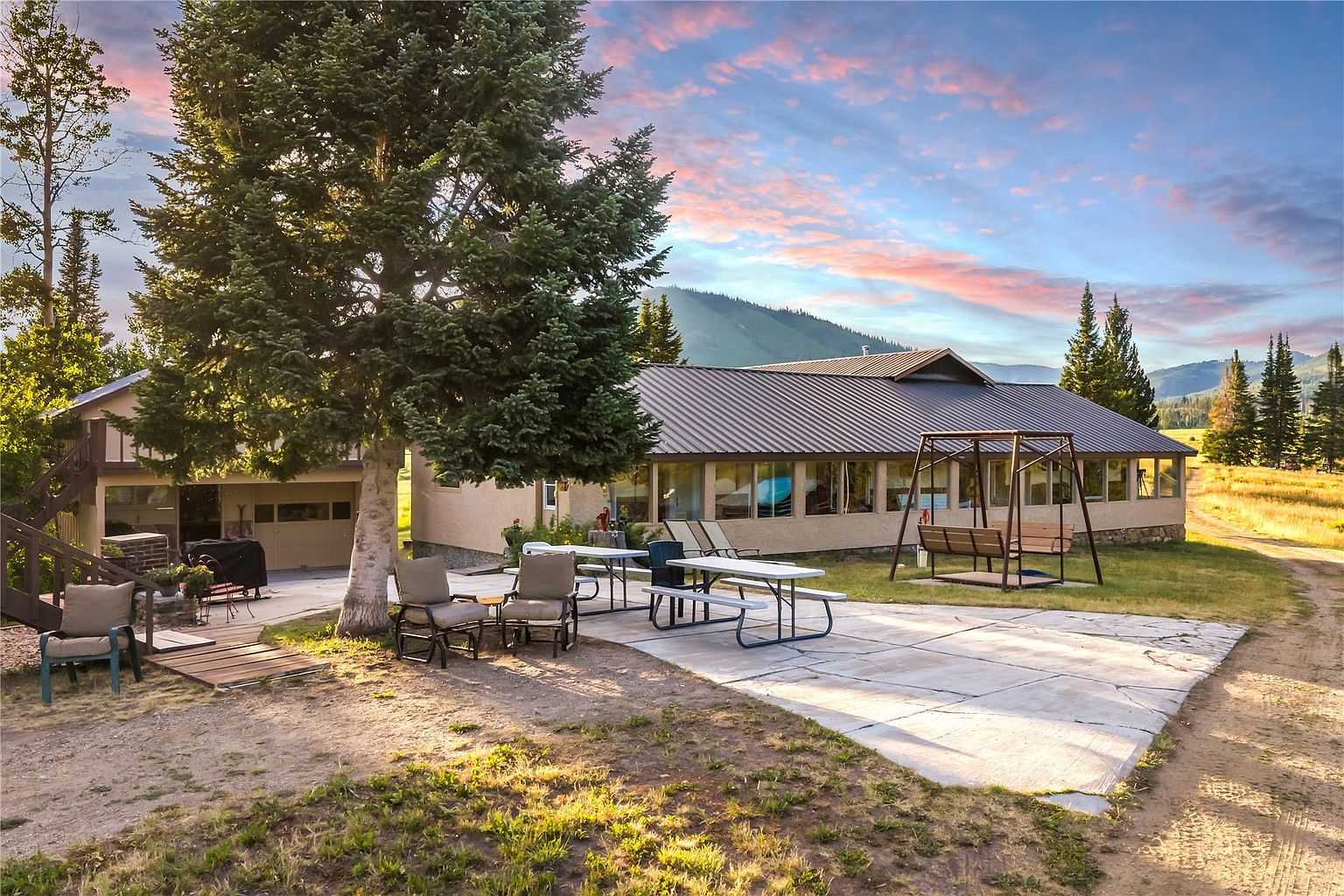 This rear exterior view showcases a sprawling single-story home with a metal roof, set against a backdrop of rolling hills and a vibrant sunset sky. The property features a large concrete patio area furnished with a picnic table, outdoor seating, and a swing, all shaded by a mature evergreen tree. The scene captures a peaceful, rural atmosphere with expansive grounds and a natural landscape.