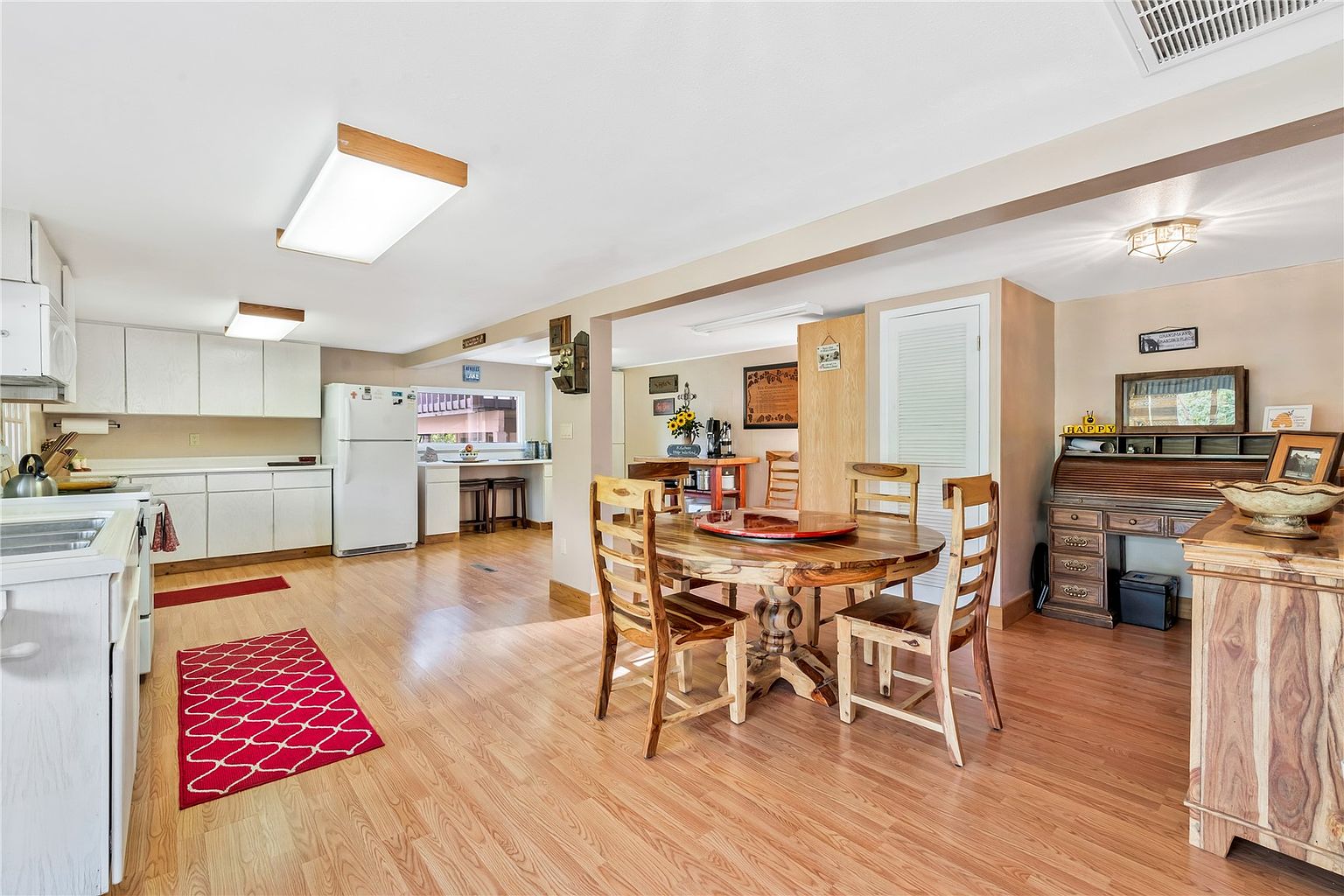 This open-concept kitchen and dining area features light wood-look flooring and white cabinetry, creating a bright and functional living space. A round wooden dining table serves as a central focal point, while a roll-top desk and decorative wall art add a touch of traditional charm to the room. The perspective captures the seamless flow between the cooking area and the dining space, highlighting the home's inviting and casual atmosphere.