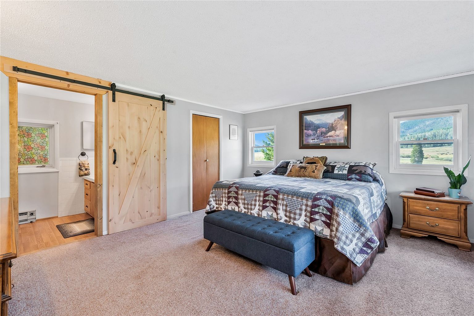 This primary bedroom features a rustic aesthetic with a large bed adorned in a patterned quilt and a blue tufted bench at the foot. A sliding barn door provides access to an adjacent bathroom, while two windows offer natural light and views of the surrounding landscape. The room is finished with neutral wall paint and carpet, creating a cozy and inviting atmosphere.
