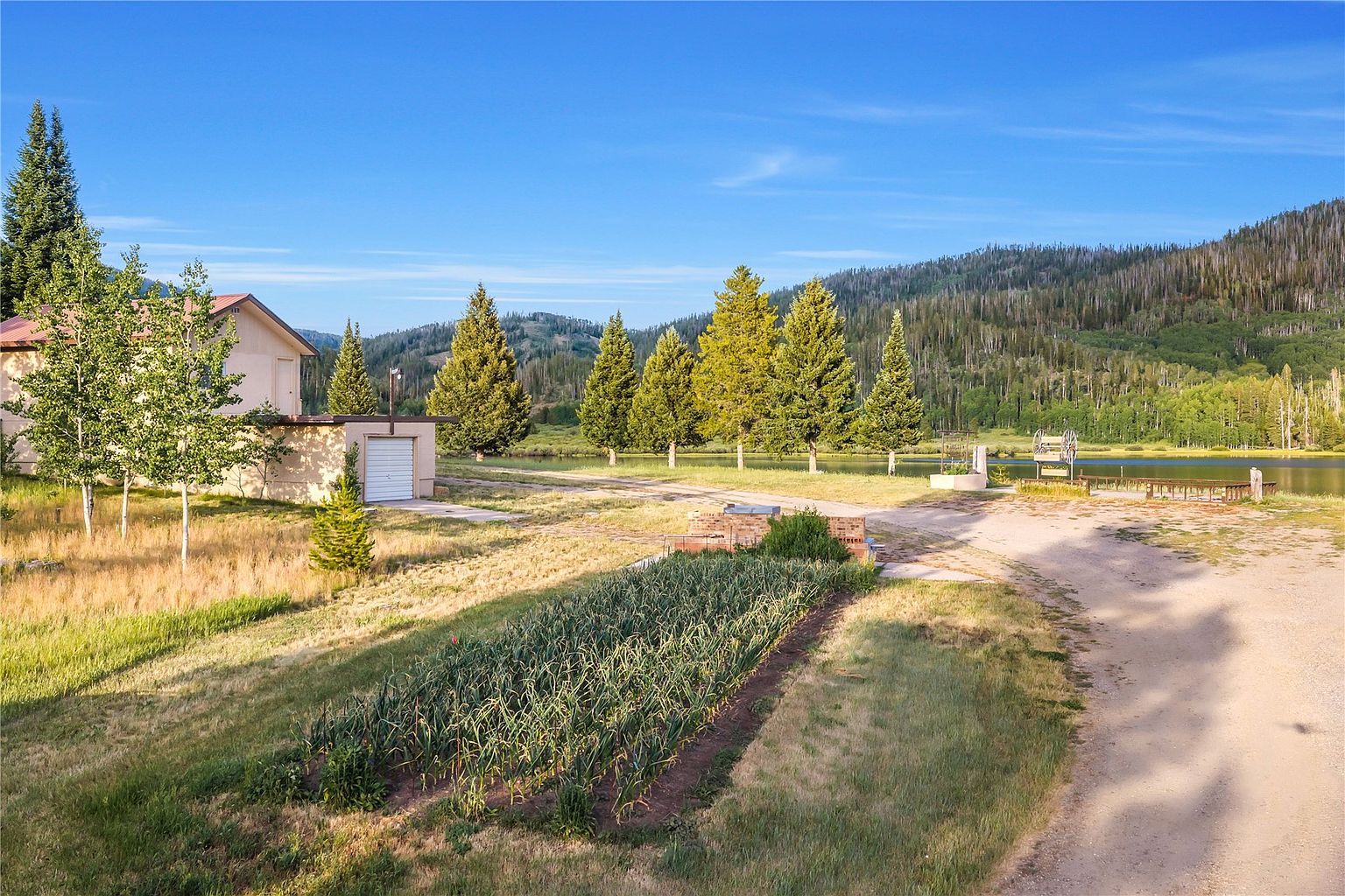 This wide-angle exterior shot captures a rural property featuring a large vegetable garden in the foreground and a detached garage building to the left. A dirt driveway leads toward a serene lake nestled against a backdrop of rolling, tree-covered mountains under a clear blue sky. The scene conveys a peaceful, expansive atmosphere, highlighting the property's integration with the natural landscape.