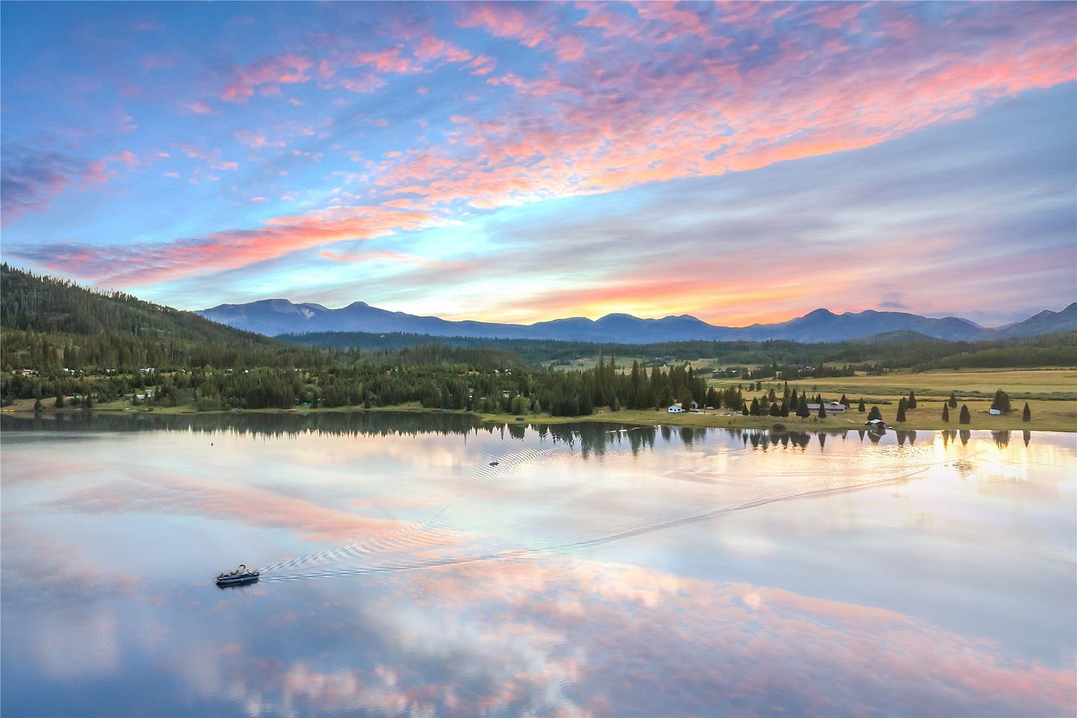 This stunning aerial view captures a serene lake at sunset, reflecting the vibrant pink and orange hues of the sky. The landscape features a dense forest of pine trees, rolling hills, and a distant mountain range, with a small boat creating a gentle wake on the calm water. The composition emphasizes the peaceful, expansive natural setting, highlighting the property's prime location near the water's edge.