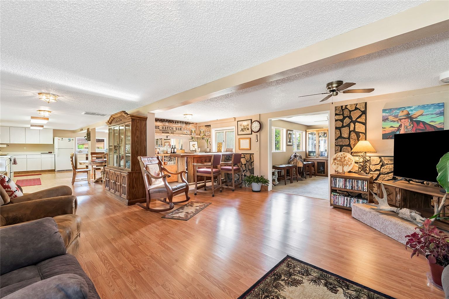This open-concept living area features warm wood-look flooring and a spacious layout that flows into a bar area and kitchen. A prominent stone-clad chimney breast anchors the living space, which includes a television, a media console, and a comfortable seating area. The room is illuminated by recessed lighting and a ceiling fan, creating a cozy, rustic atmosphere with a clear view into the adjacent dining and kitchen zones.