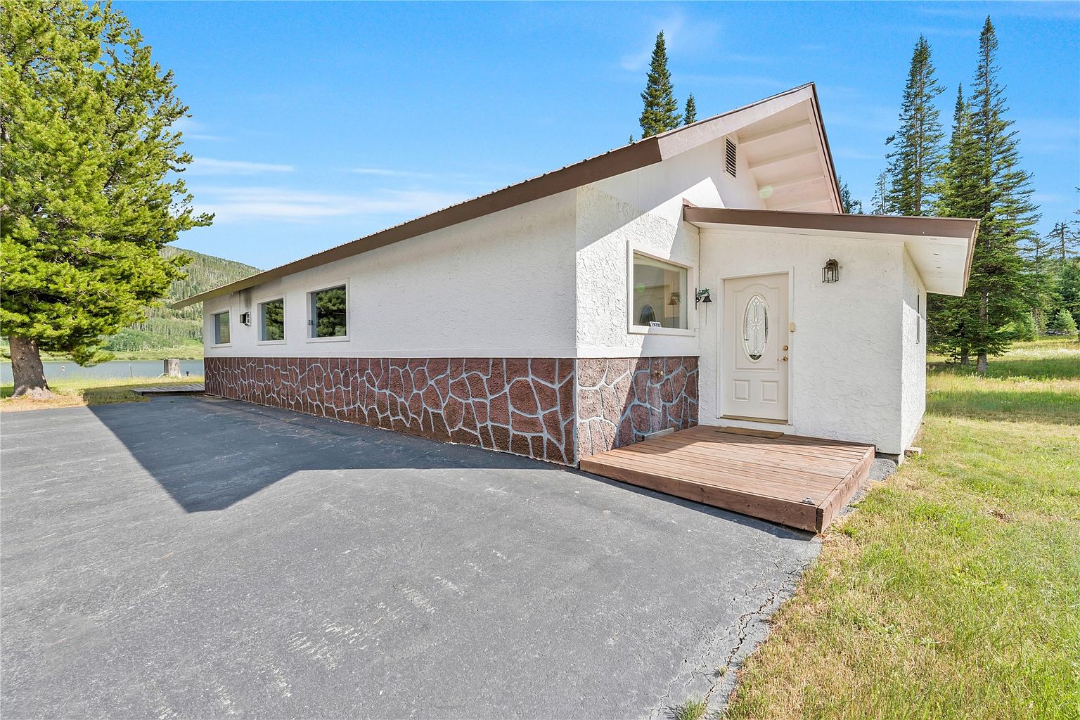 This exterior view captures the front entrance of a single-story home featuring a white stucco finish accented by a decorative stone-clad base. A wooden deck leads to a white front door with a decorative glass insert, while a paved driveway extends along the side of the house. The property is situated in a scenic, wooded environment with tall pine trees and a glimpse of a nearby lake in the background.