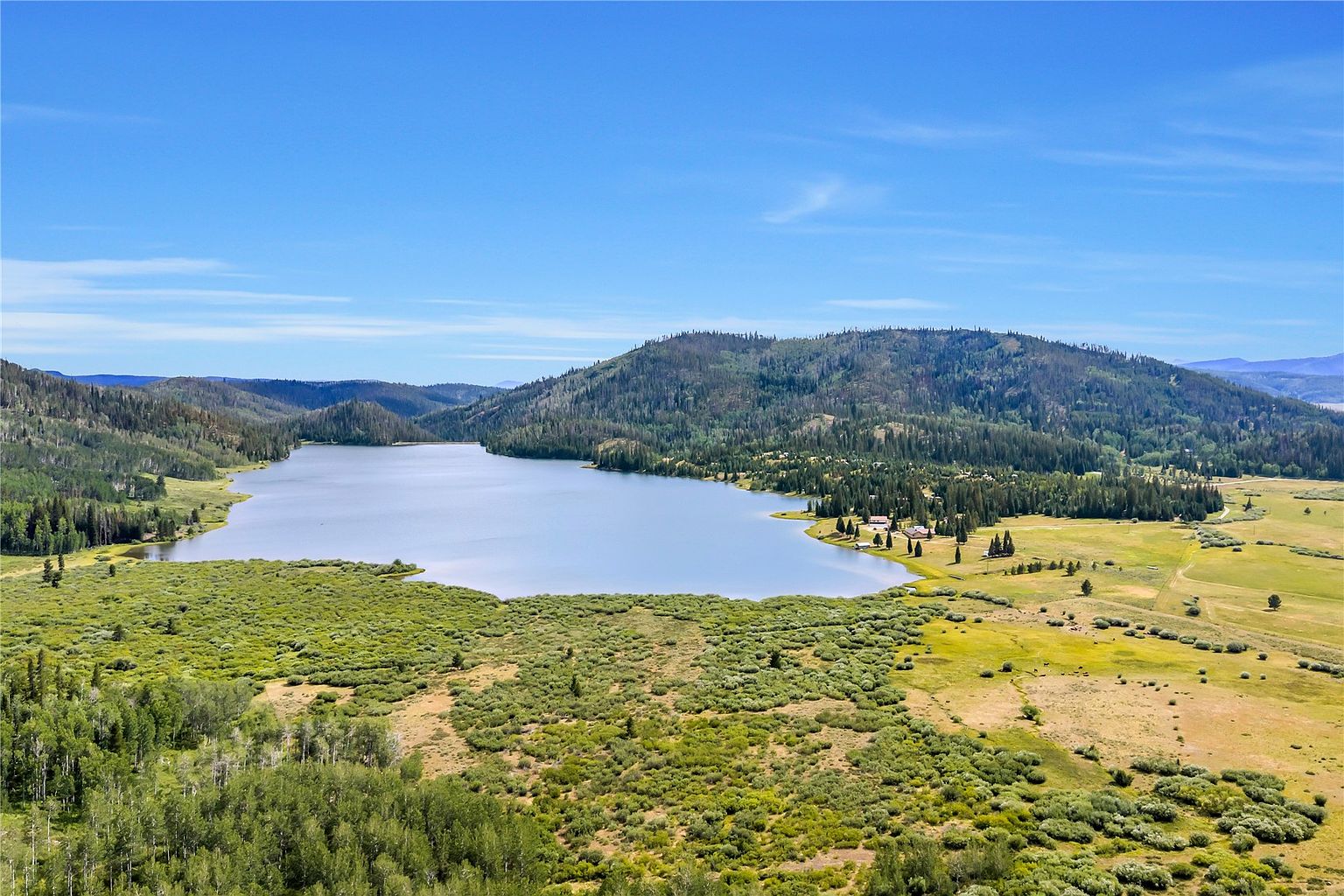 This expansive aerial view captures a serene mountain lake surrounded by lush green forests and rolling meadows. The perspective highlights the vast natural landscape, showcasing the property's secluded location and proximity to the water. The clear blue sky and vibrant greenery create a peaceful and picturesque setting, emphasizing the property's potential for outdoor recreation and tranquility.