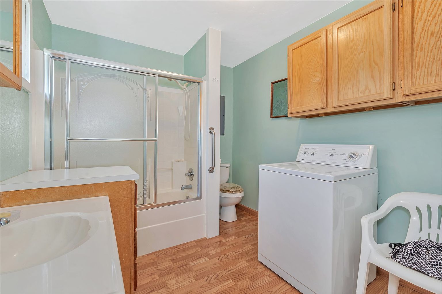 This multi-functional space combines a laundry area with a bathroom, featuring light green walls and wood-look flooring. The room includes a white top-load washing machine positioned beneath light wood wall cabinets, a shower stall with a glass enclosure, and a toilet tucked into a corner. A white plastic chair sits in the foreground, providing a practical seating option in this utilitarian room.