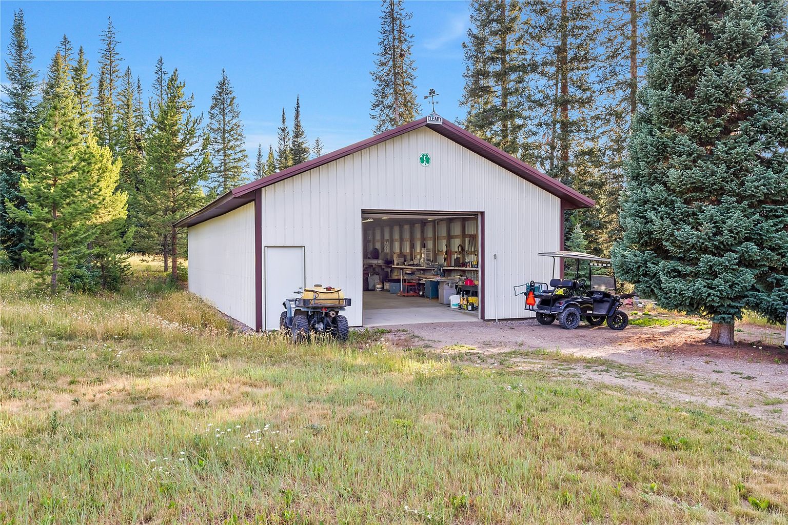 This image features a detached, metal-sided workshop or garage building set in a wooded, rural landscape. The structure has a large, open bay entrance revealing a well-organized interior workspace, with an ATV parked to the left and a golf cart to the right. The building is topped with a weather vane and surrounded by tall pine trees, creating a quiet, functional outdoor setting.