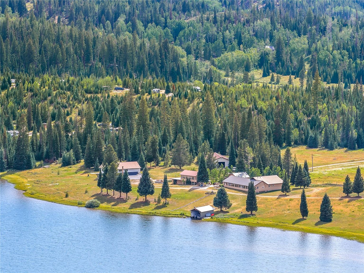 This high-angle aerial view captures a sprawling rural property situated directly along the edge of a serene lake. The landscape features a collection of residential buildings and outbuildings nestled among dense evergreen forests and open grassy meadows. A small boat house sits at the water's edge, emphasizing the property's direct access to the lake and its tranquil, secluded setting.