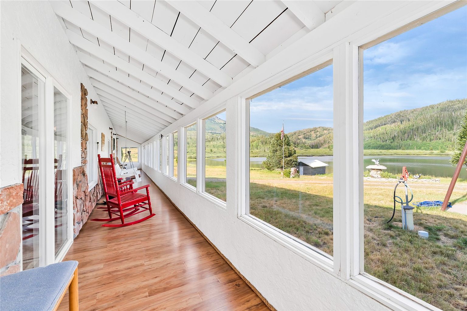 This bright sunroom features a long row of large windows that provide expansive views of a lake and surrounding mountains. The space is furnished with vibrant red rocking chairs and has light wood-look flooring, creating a cozy and inviting atmosphere. A stone-clad wall adds a rustic texture to the interior, while the white-painted exposed beam ceiling enhances the airy, open feel of the room.
