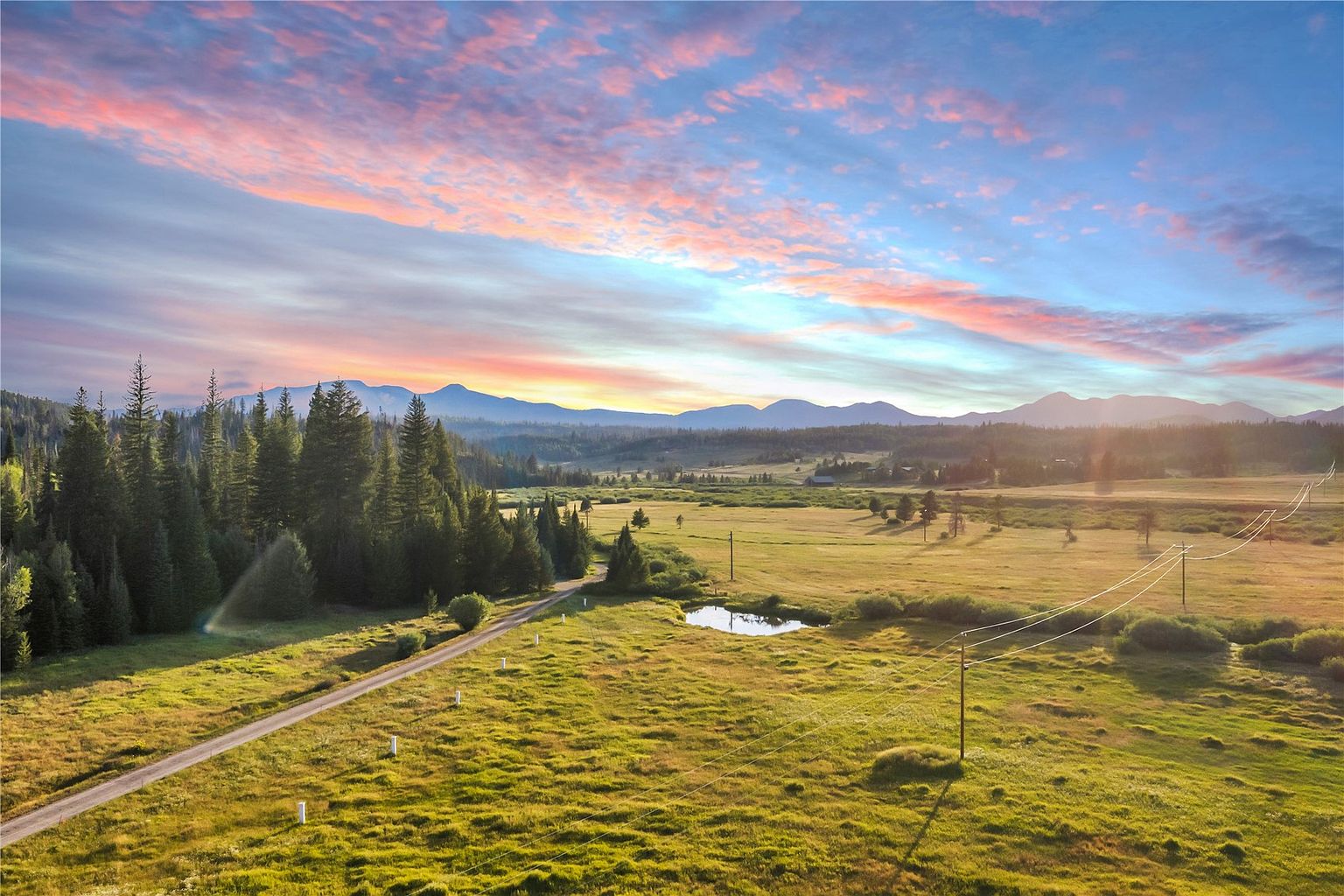 This expansive aerial view captures a vast, rolling meadow landscape set against a backdrop of majestic mountain ranges under a vibrant, colorful sunset sky. A gravel road winds through the grassy terrain, leading toward a small pond, while utility lines stretch across the foreground, emphasizing the scale of the property. The scene conveys a sense of serene, wide-open space and natural beauty, ideal for those seeking a secluded rural retreat.