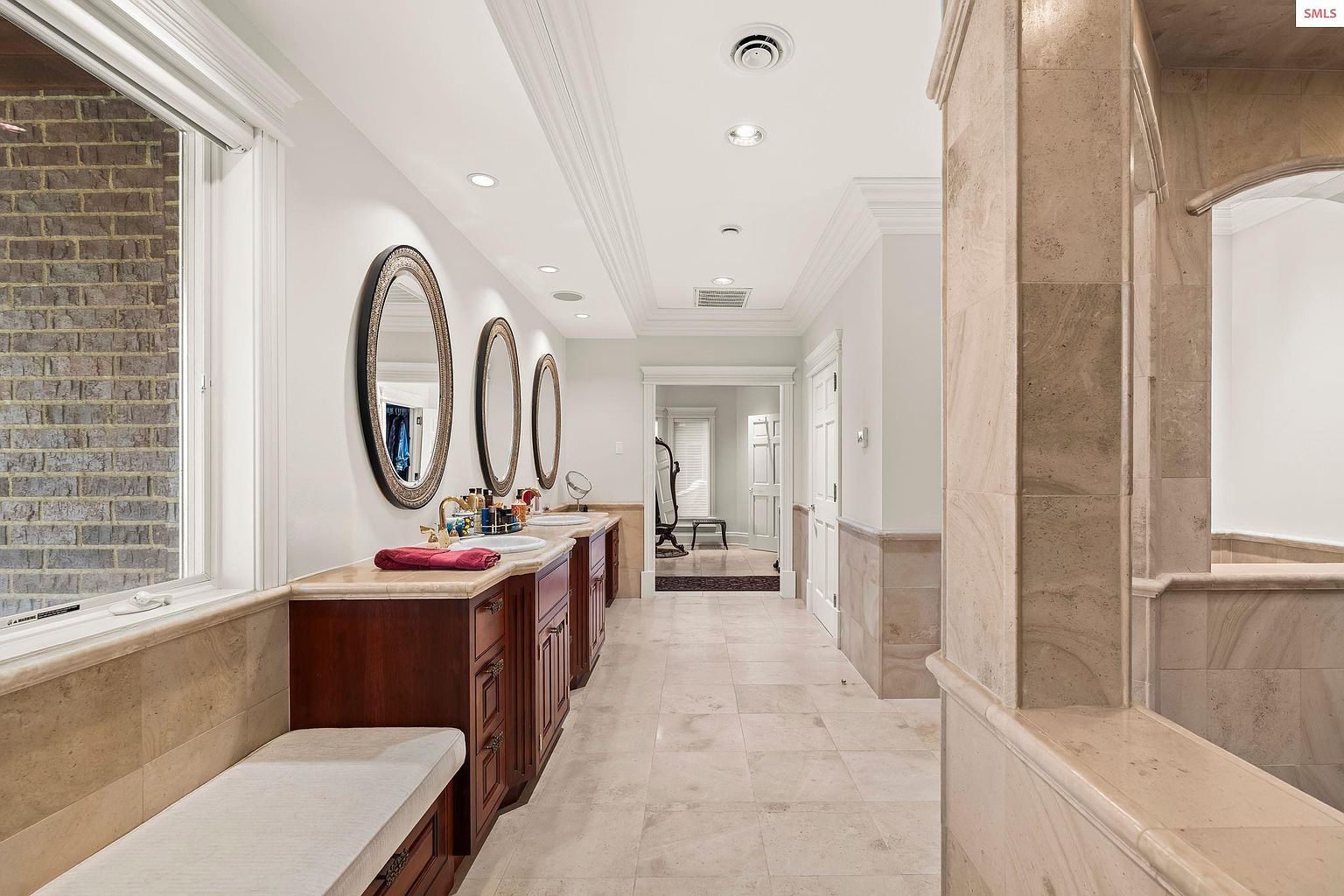This is a luxurious primary bathroom featuring a long vanity with dual sinks, framed oval mirrors, and dark wood cabinetry. A built-in bench sits beneath a window, and the floor and walls are covered in light-colored stone tiles. The bathroom leads to another area through an arched opening, creating a spa-like atmosphere.