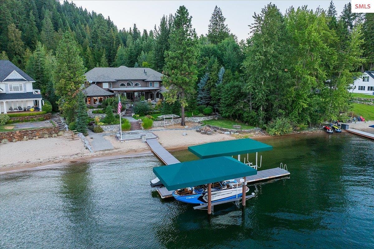 This aerial view showcases a stunning waterfront property featuring a large house with a well-manicured lawn and mature trees. The property includes a private beach, a dock with boat lifts, and clear water, creating an idyllic lakeside setting. The overall impression is one of luxury and tranquility.