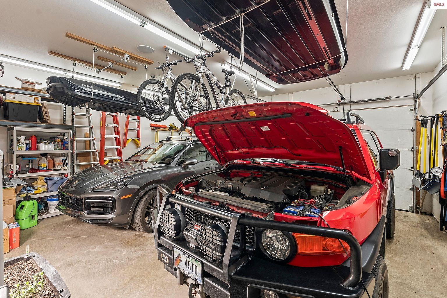 This is a well-organized garage featuring two cars, a red Toyota FJ Cruiser with its hood open and a gray Porsche Cayenne. Overhead storage solutions include bike racks and cargo carriers, maximizing space. The garage is brightly lit with fluorescent lighting, and shelving units provide additional storage for tools and supplies.