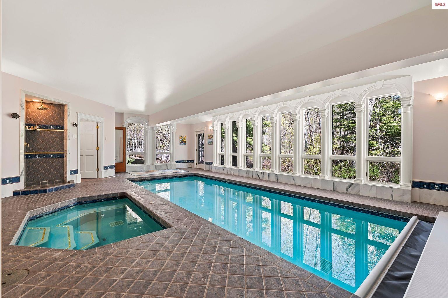 This image showcases an indoor pool and spa area, featuring a rectangular pool with clear blue water and a separate hot tub. Large windows line one side of the room, providing natural light and views of the surrounding trees. The tiled flooring and architectural details, such as the arched window frames, add a touch of elegance to the space.