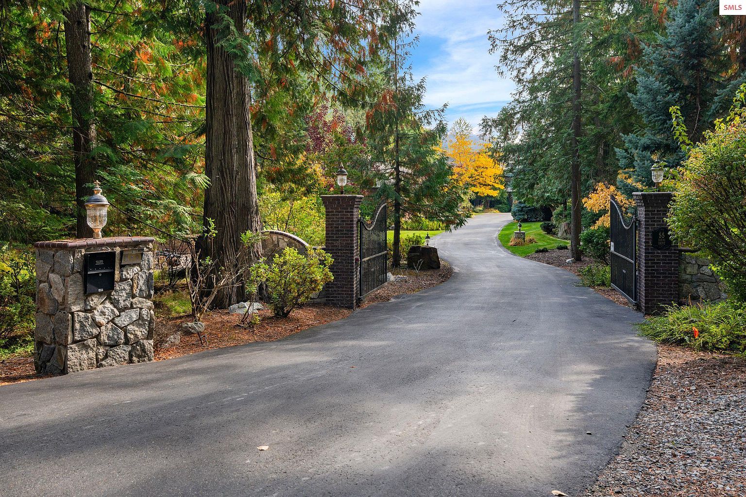 The image showcases a grand entryway to a property, featuring a long, winding asphalt driveway flanked by mature trees with autumn foliage. Stone and brick pillars support ornate metal gates on either side of the entrance, with decorative lanterns atop the pillars. The scene evokes a sense of privacy and upscale living.