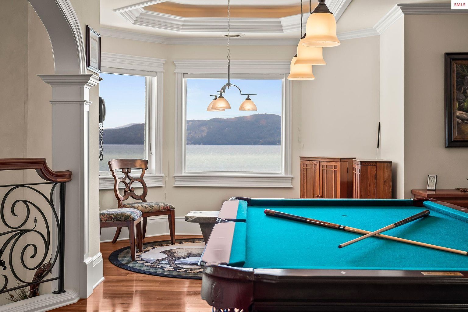 This interior shot showcases a well-appointed living room featuring a pool table with cues, hardwood floors, and large windows offering a scenic view of a lake and mountains. The room is illuminated by pendant lighting, and a decorative chair and rug add to the room's character. The overall impression is one of luxury and relaxation.