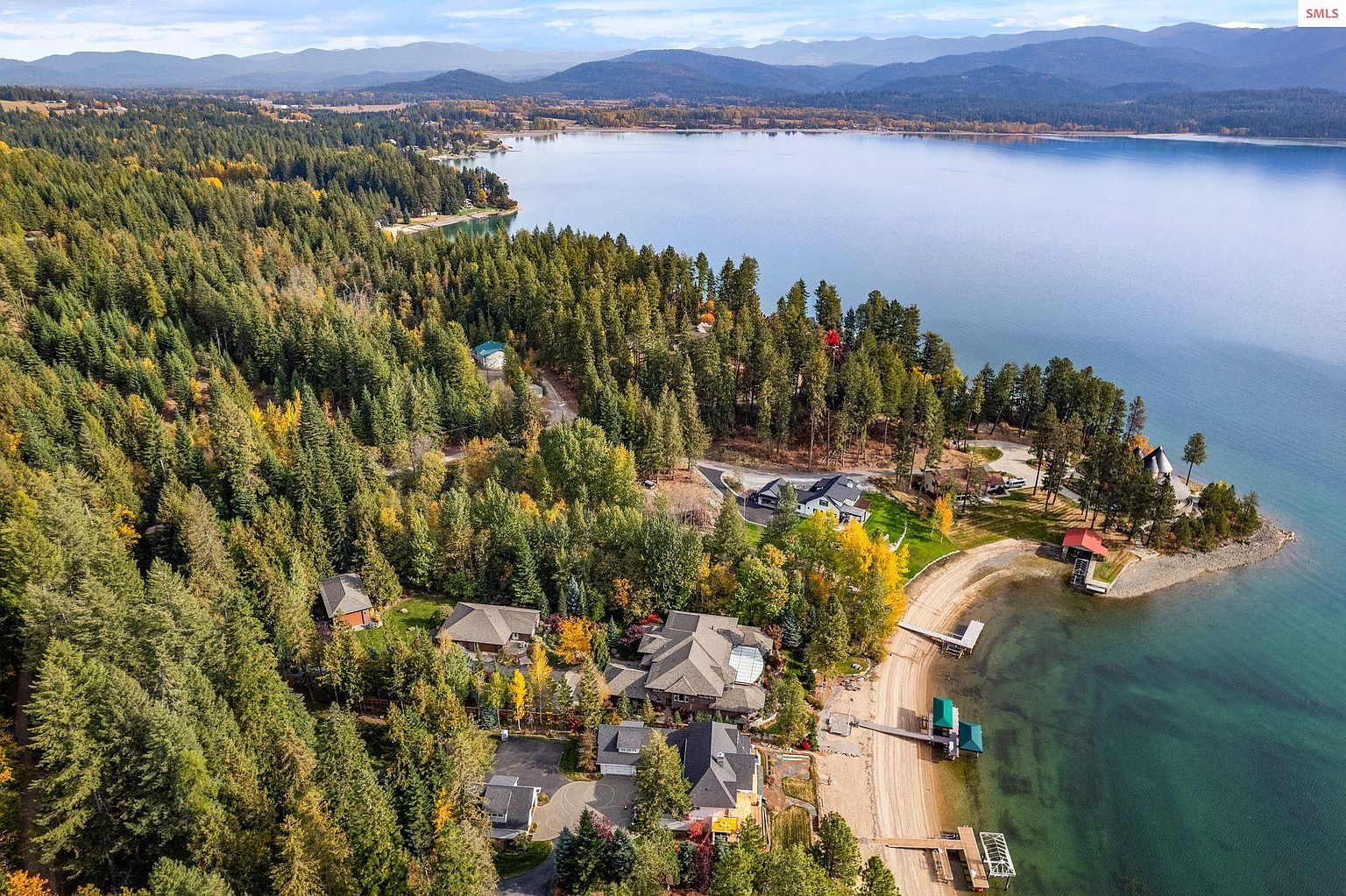 This aerial view showcases a luxurious lakeside property surrounded by lush forests and mountains. The homes feature architectural details and private docks, offering direct access to the pristine lake. The landscape is a mix of evergreens and deciduous trees, creating a picturesque and serene setting.