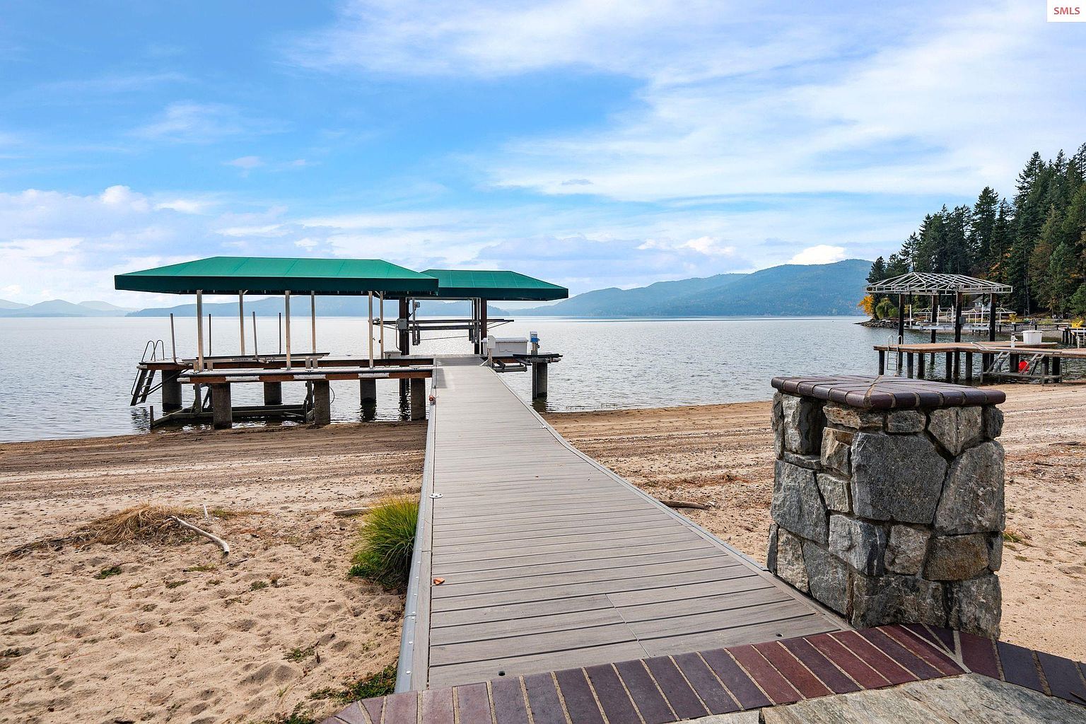 This exterior shot showcases a beautiful lakeside property featuring a long wooden dock leading to covered boat slips. The dock is flanked by sandy beaches and a stone pillar, adding a touch of rustic charm. In the background, mountains and lush greenery create a serene and picturesque setting, perfect for waterfront living.