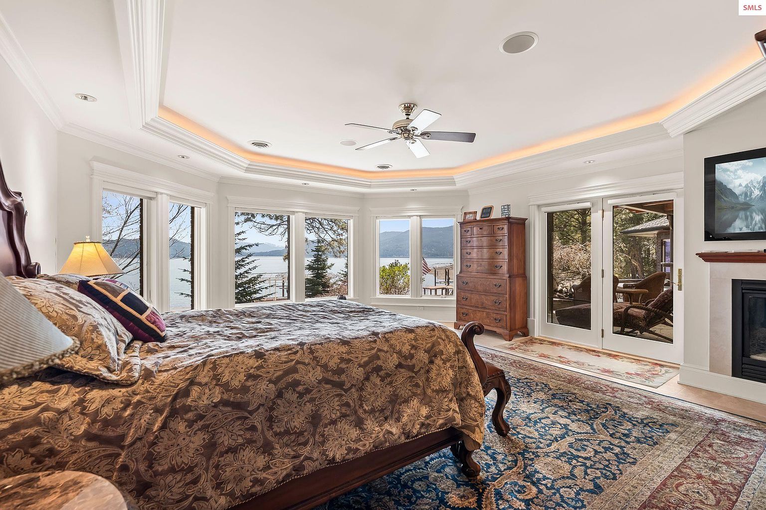 This is a primary bedroom featuring a large bed with a patterned comforter, a tall wooden dresser, and a view of a lake through multiple windows. The room has a tray ceiling with accent lighting and a ceiling fan. A fireplace and television are visible on the right side of the image, and a patterned rug covers the floor, adding warmth and texture to the space.