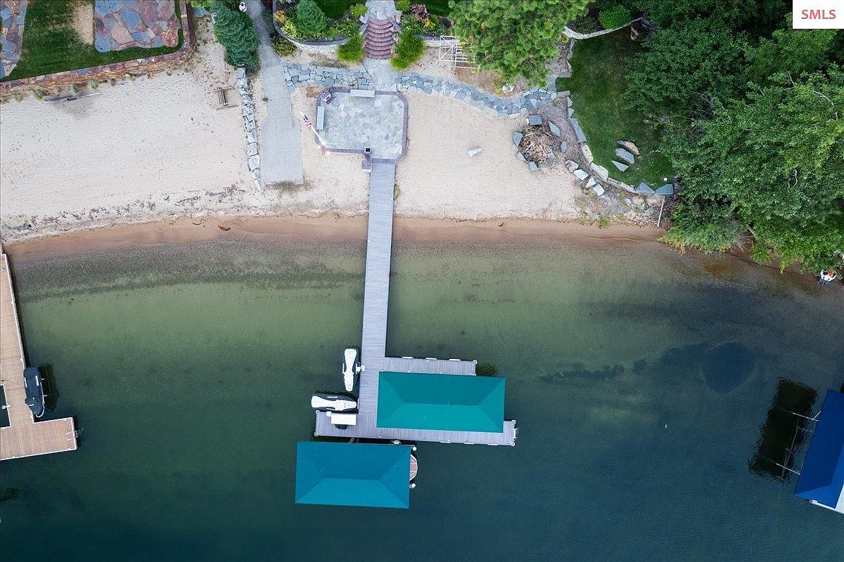 This aerial view showcases a stunning waterfront property with a private beach, a long wooden pier, and boat docks featuring green coverings. The landscape includes manicured lawns, mature trees, and stone pathways leading to the water. The overall impression is one of luxury and serene lakeside living.