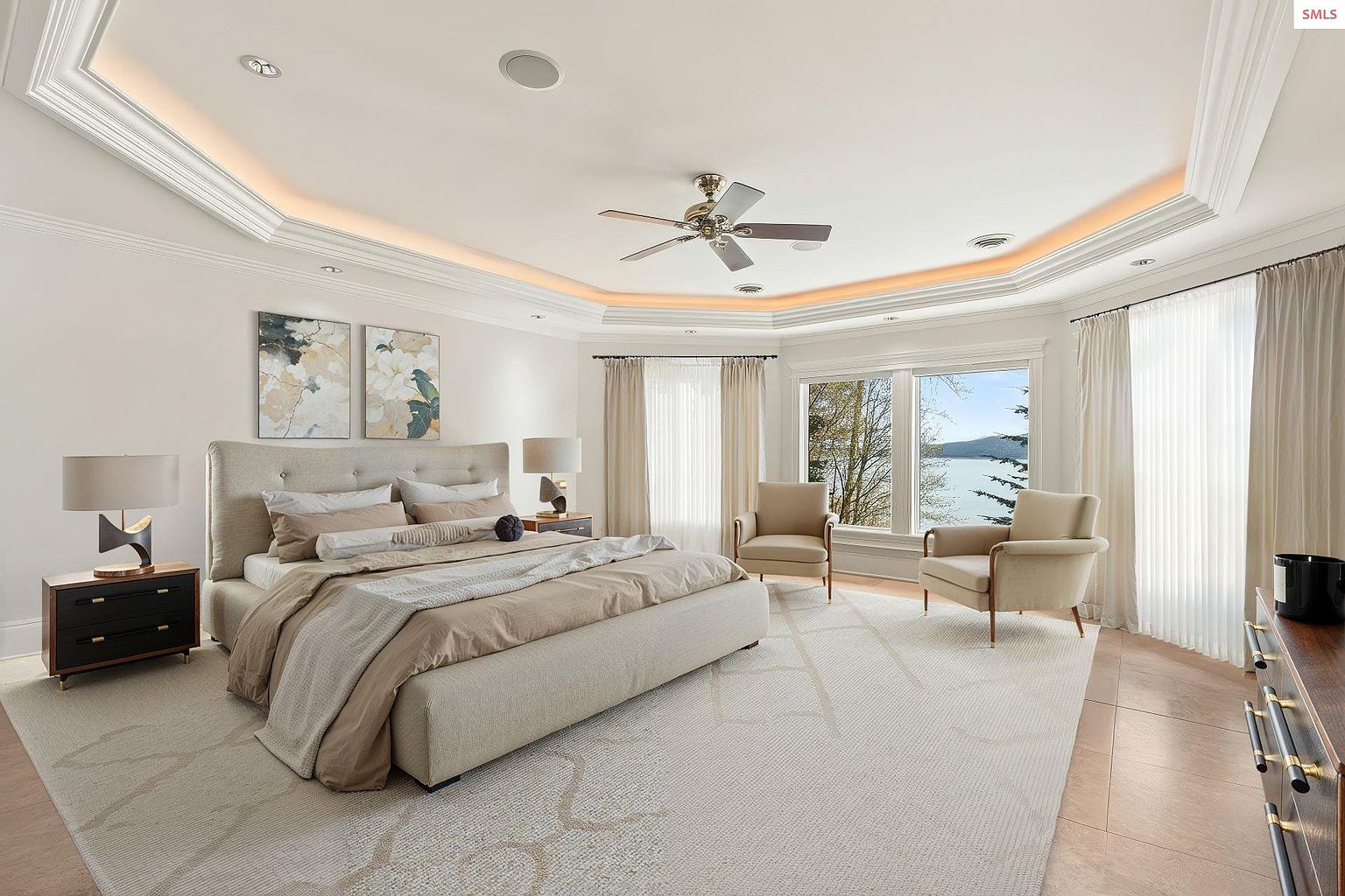 This is a luxurious primary bedroom featuring a large bed with neutral-toned bedding, flanked by matching nightstands and lamps. The room has a tray ceiling with accent lighting, a ceiling fan, and large windows with sheer curtains that offer a view of the water. Two armchairs are placed near the windows, creating a cozy seating area.