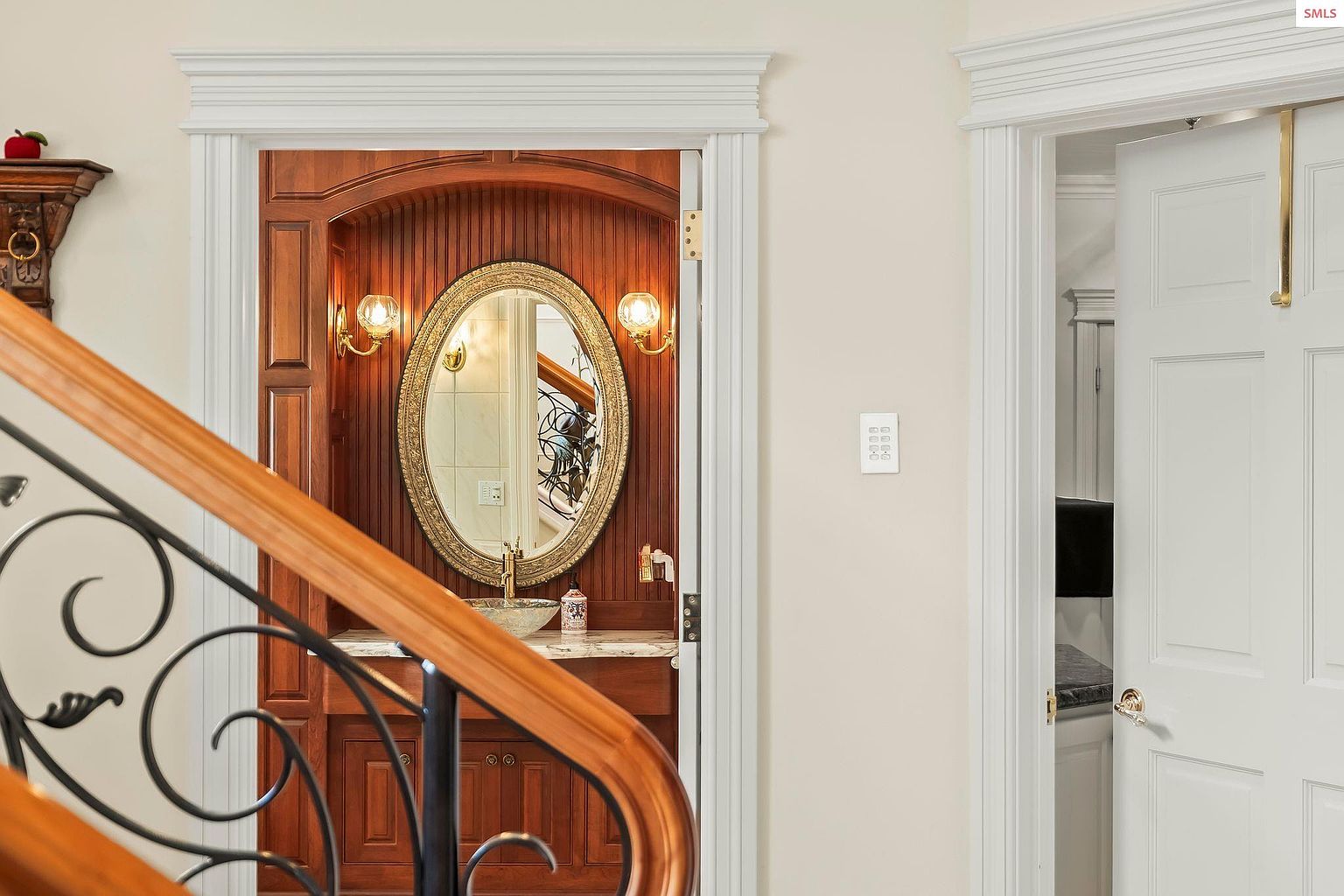 This image showcases a hallway with a glimpse into a well-appointed powder room. The powder room features rich wood paneling, an oval mirror with gold accents, and sconce lighting. The hallway includes a staircase with a decorative wrought iron railing and a doorway leading to another room, creating an elegant and inviting transition space.