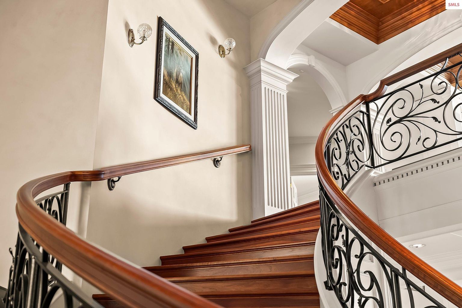 This interior shot showcases a beautifully designed staircase with wooden steps and an ornate wrought iron railing. The curved design adds a touch of elegance, complemented by the warm wood tones and the natural light filtering through. A framed picture and sconces adorn the wall, enhancing the sophisticated ambiance.
