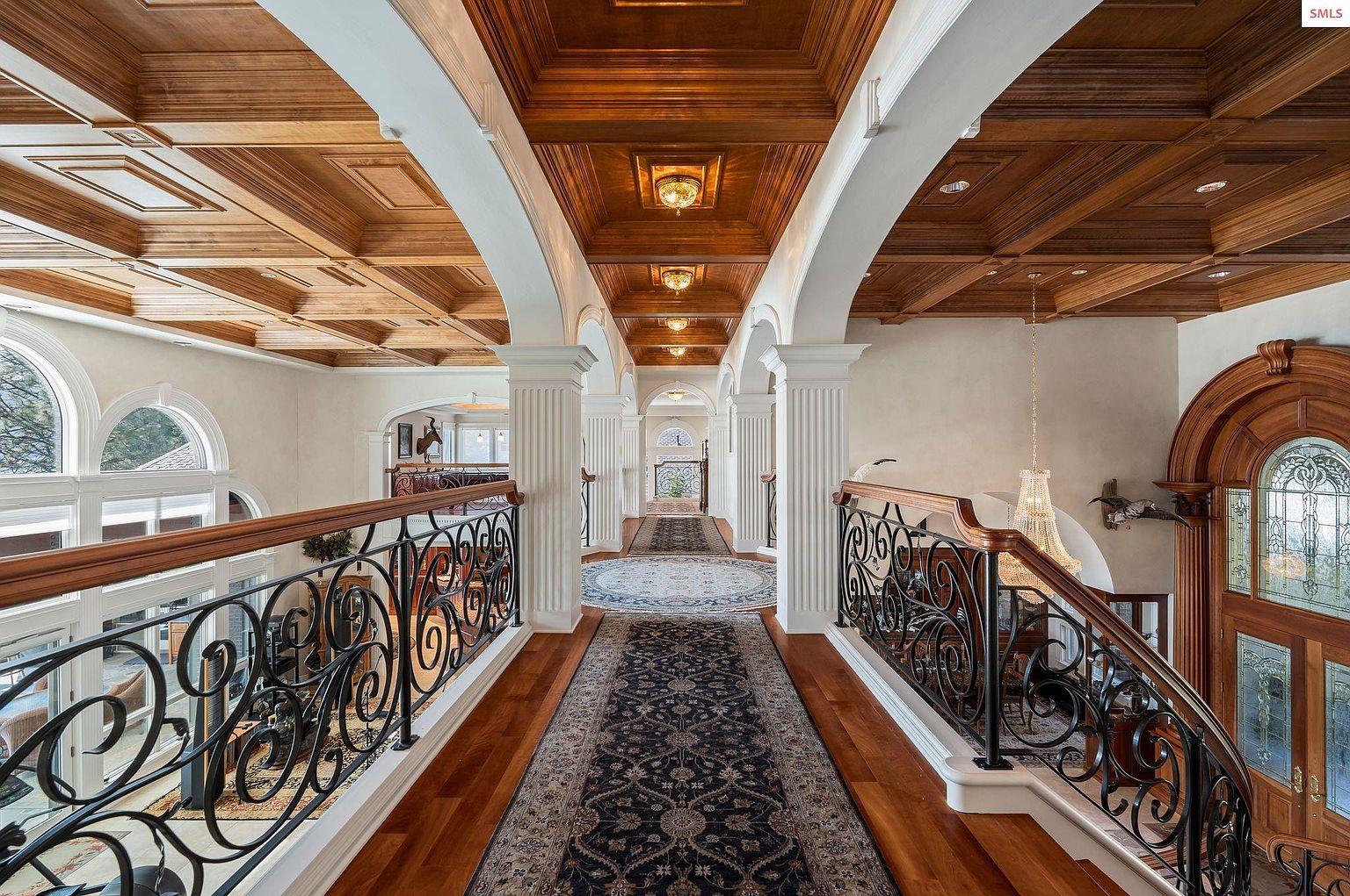 This is an interior shot of a grand hallway featuring a coffered wood ceiling, white columns, and ornate wrought iron railings. A long patterned runner stretches down the hardwood floor, leading towards a distant doorway. The space exudes elegance and sophistication, suggesting a high-end property.