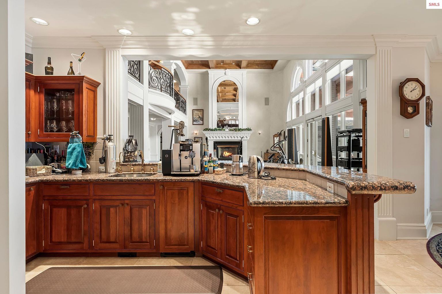 This interior shot showcases a well-appointed kitchen area with rich, dark wood cabinetry and granite countertops. The space opens up to a grand living area with a fireplace and large windows, creating a sense of spaciousness and luxury. A coffee station is prominently featured, suggesting a focus on convenience and modern living.
