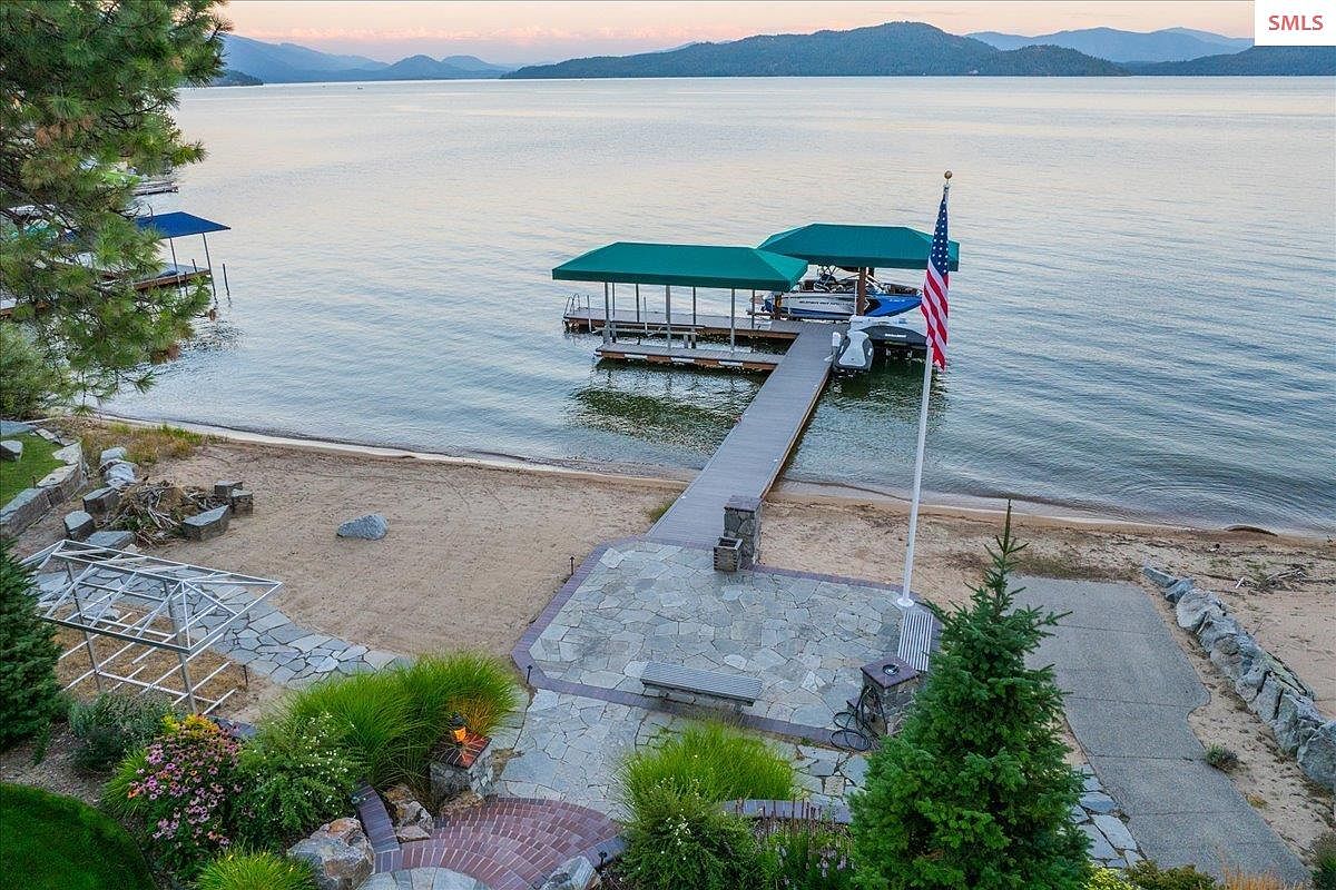 This aerial view showcases a stunning waterfront property with a private dock and boat lift. The property features a well-maintained yard with mature landscaping, a stone patio, and a sandy beach area. The American flag waves proudly near the dock, adding a touch of patriotism to this idyllic lakeside setting.