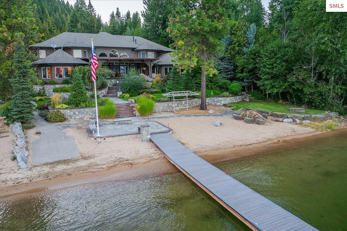 This is an exterior rear view of a luxurious waterfront property. The house features a multi-level landscaped yard leading down to a private beach with a dock extending into the water. The architecture includes a mix of stone and brick elements, with large windows and a covered patio area, creating an inviting outdoor living space.