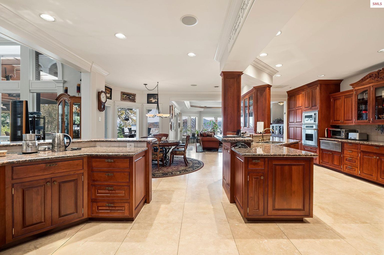 This is a spacious kitchen with warm wood cabinetry, granite countertops, and tile flooring. The kitchen features two islands, stainless steel appliances, and an open layout that connects to the dining and living areas. Large windows provide ample natural light, enhancing the inviting atmosphere.