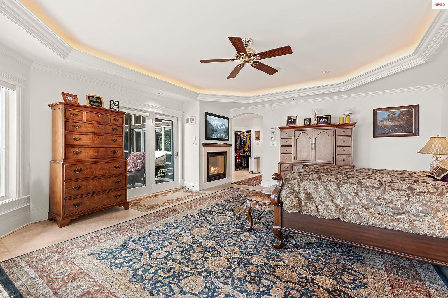 This is a primary bedroom featuring a large bed with a patterned comforter, a fireplace with a television above it, and a ceiling fan. The room has a luxurious feel with detailed crown molding, a large area rug, and a walk-in closet visible through an archway. The perspective is from the foot of the bed, showcasing the room's depth and features.