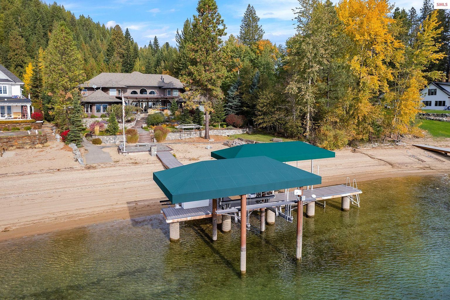 This aerial view showcases a stunning lakeside property featuring a large, multi-story house nestled among mature trees. A private dock with boat covers extends into the clear water, complemented by a sandy beach. The landscape includes well-maintained gardens and lush greenery, creating a serene and luxurious atmosphere.