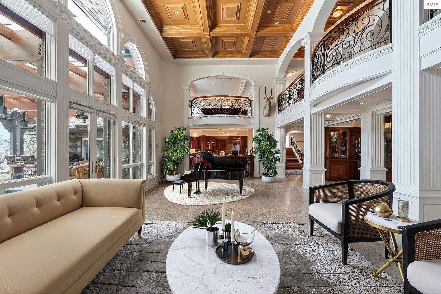 This grand living room features soaring ceilings with a coffered wood design, large windows providing ample natural light, and a balcony overlooking the space. A black grand piano sits on a circular rug, complemented by elegant furniture and decor, creating a luxurious and inviting atmosphere. The perspective is from the front of the room, showcasing the depth and grandeur of the space.