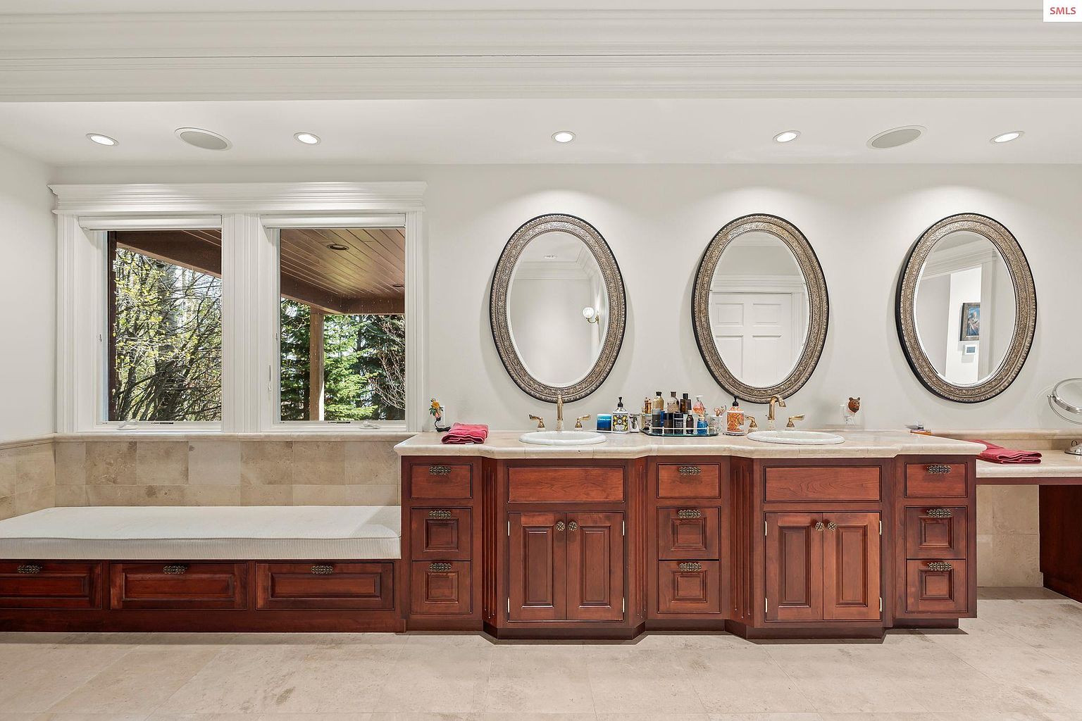 This is a primary bathroom featuring a double vanity with dark wood cabinetry and oval mirrors. The countertop is light-colored stone, and there is a built-in bench seat under a window. The overall style is traditional and luxurious, with a focus on natural light and ample space.