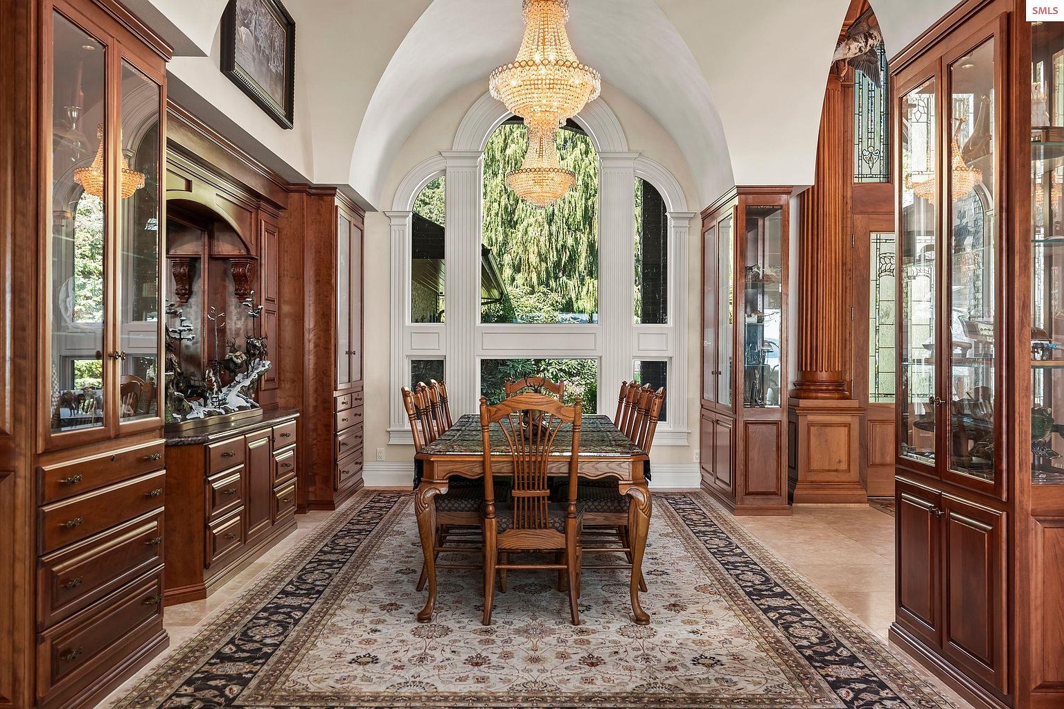 This is a formal dining room featuring a large wooden table with chairs, surrounded by built-in wooden cabinets and display cases. A grand chandelier hangs above the table, and a large arched window provides natural light and a view of the outdoors. The room is decorated with a patterned rug, adding to the elegant and traditional style.