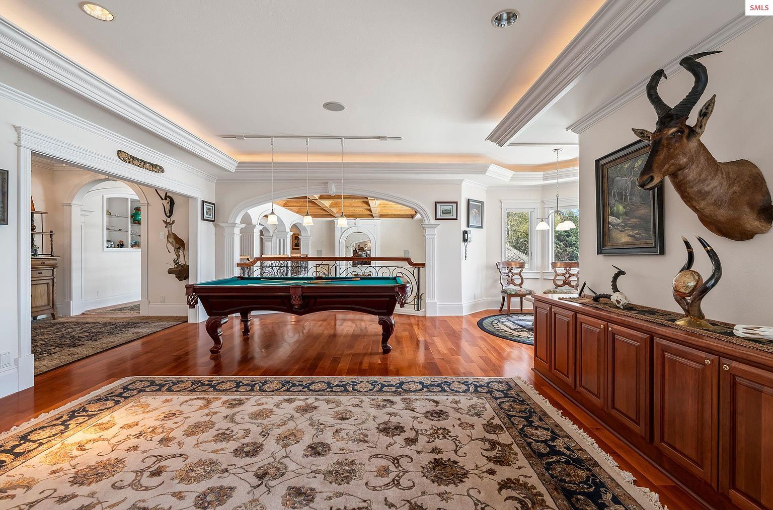 This interior shot showcases a spacious living room featuring a billiard table as a central element. The room is adorned with elegant architectural details such as arched doorways, crown molding, and recessed lighting. A large area rug anchors the space, while mounted taxidermy and artwork add character and sophistication.