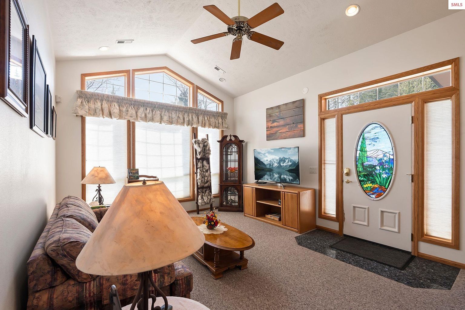 This is a cozy living room featuring a large window with a valance, a ceiling fan, and a television. The room is furnished with a patterned sofa, a coffee table, and a lamp. The front door has a decorative stained glass window and sidelights, adding a unique touch to the space.