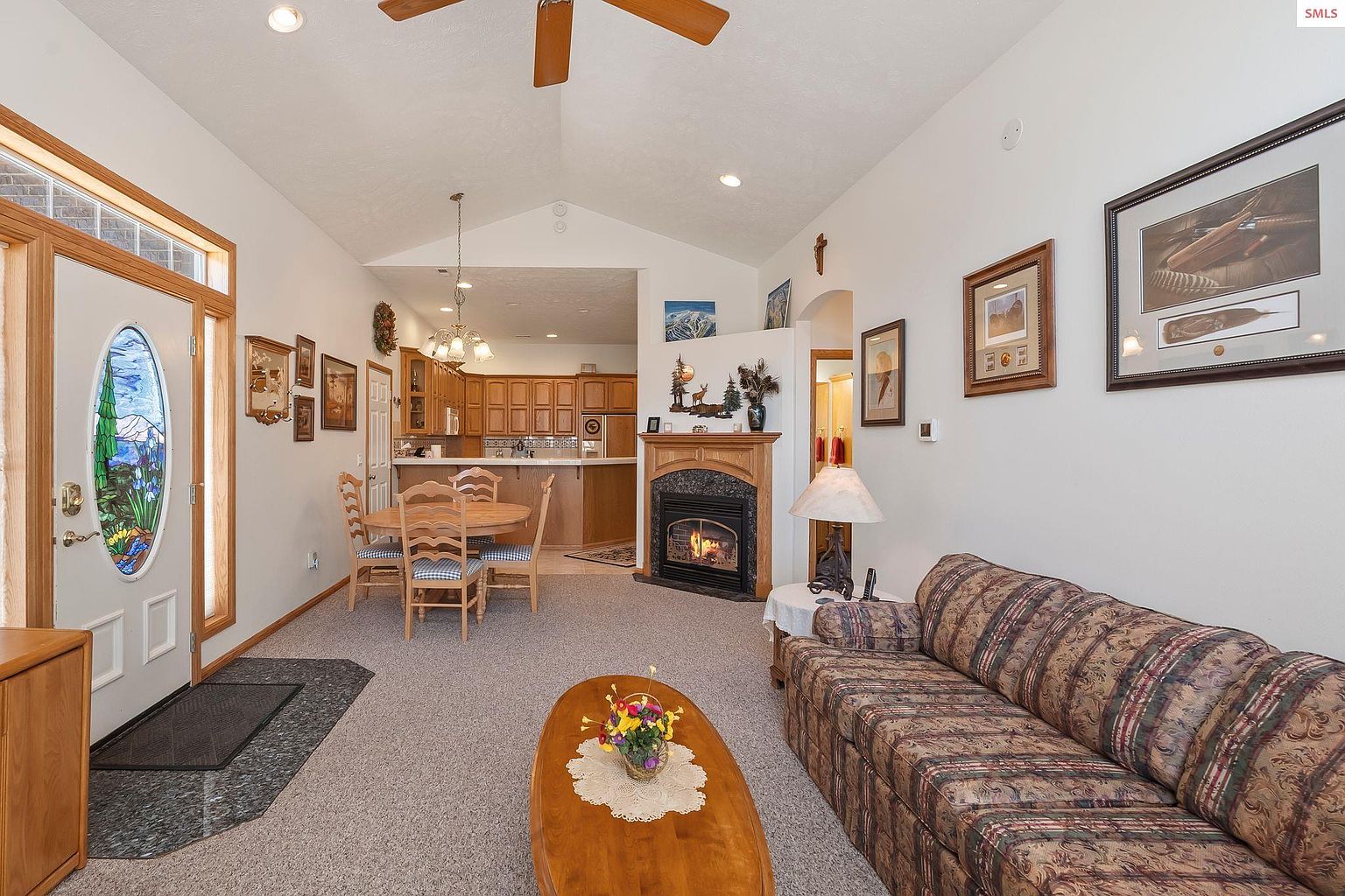 This interior shot showcases a cozy living room with a fireplace, a patterned sofa, and a wooden coffee table adorned with flowers. The room features a dining area adjacent to an open kitchen, creating a welcoming and connected living space. Natural light filters through the front door, enhancing the warm and inviting atmosphere.