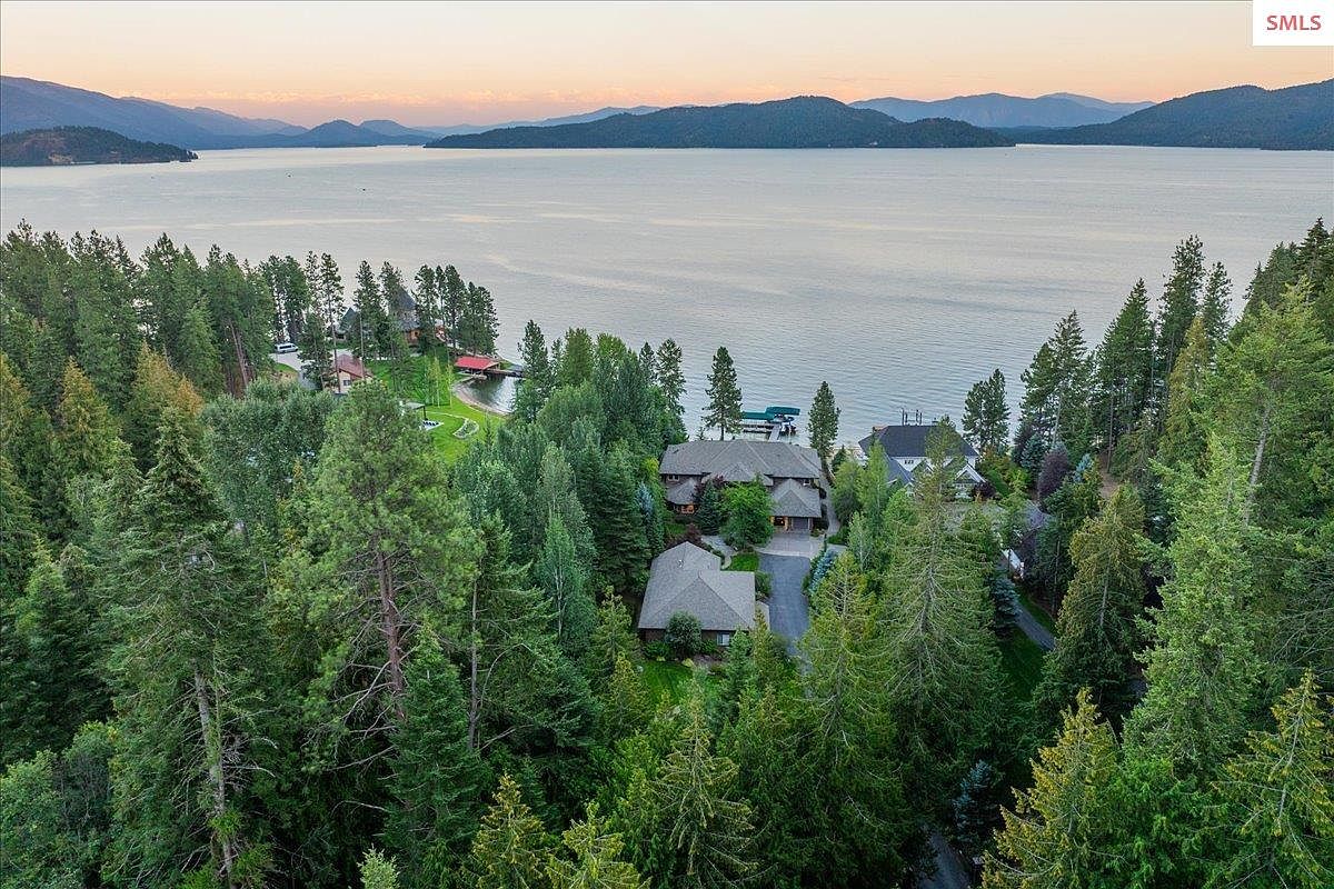 This aerial view showcases a luxurious property nestled among lush green trees, offering stunning views of a serene lake and distant mountains. The homes feature elegant architecture with well-maintained roofs and landscaping, suggesting privacy and exclusivity. The overall impression is one of tranquility and high-end living in a natural setting.