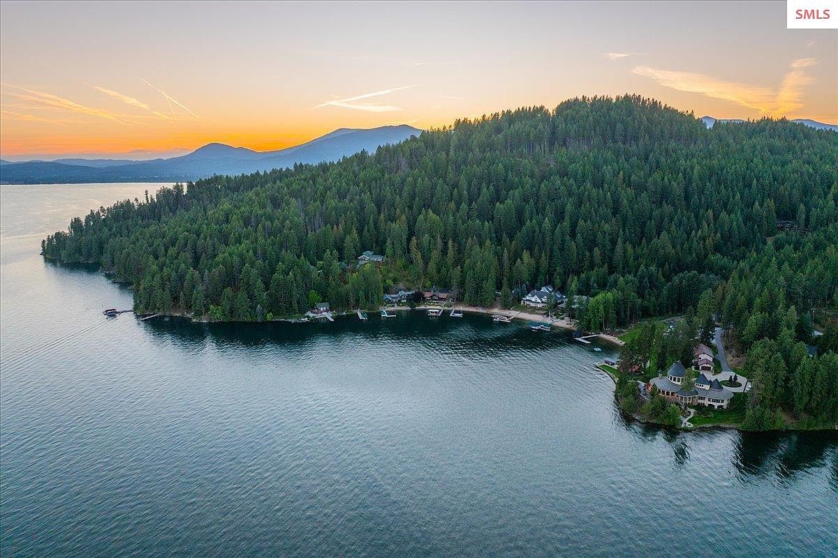 This aerial view showcases a stunning waterfront property nestled on a lush, forested peninsula. The large, luxurious home features intricate architectural details and is surrounded by mature trees, offering privacy and seclusion. The calm, reflective water and the backdrop of distant mountains create a serene and picturesque setting.