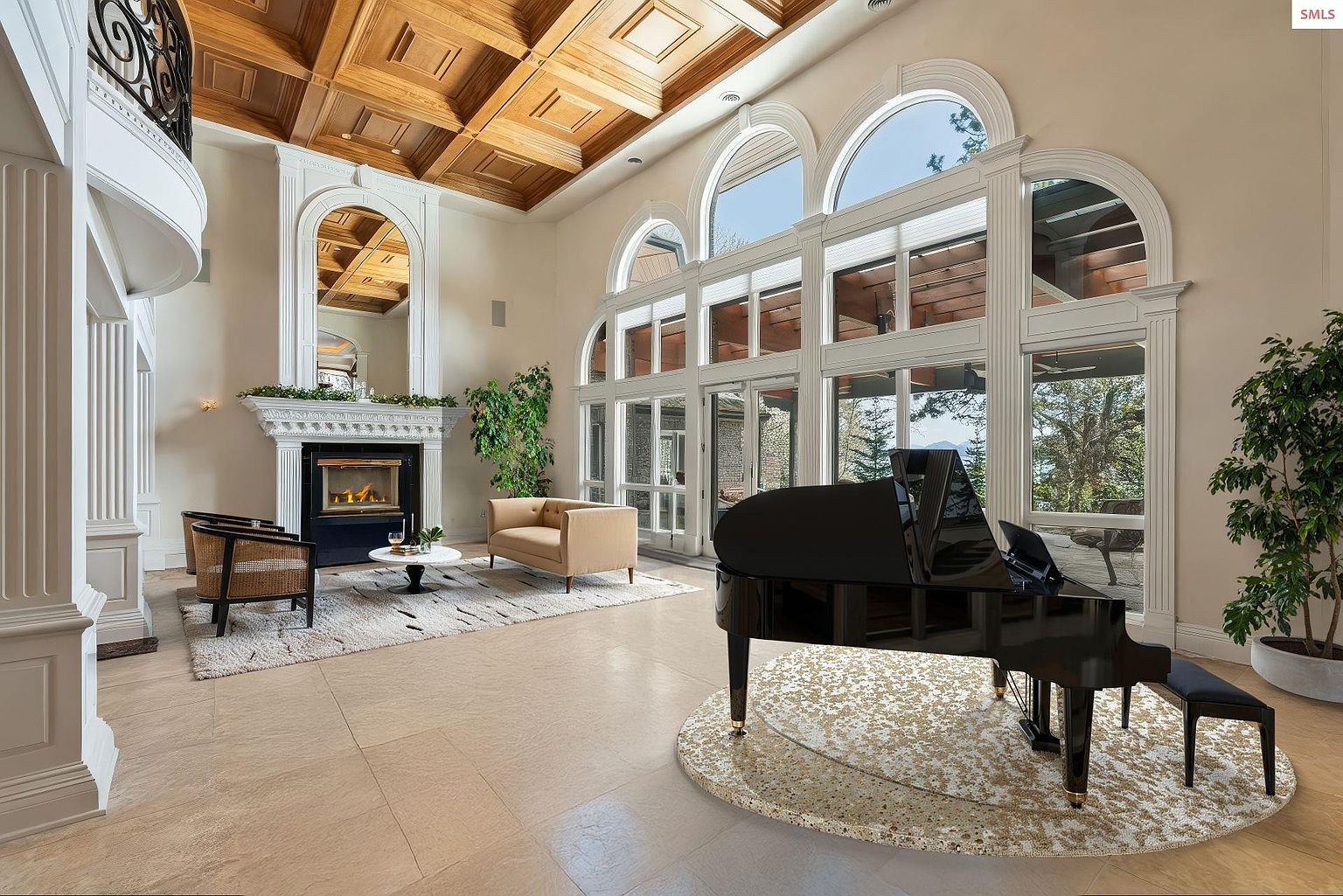 This grand living room features a coffered wood ceiling, a large arched window wall, and a fireplace with a decorative mantel. A black grand piano sits on a circular rug, adding a touch of elegance. The room is spacious and well-lit, creating a luxurious and inviting atmosphere.