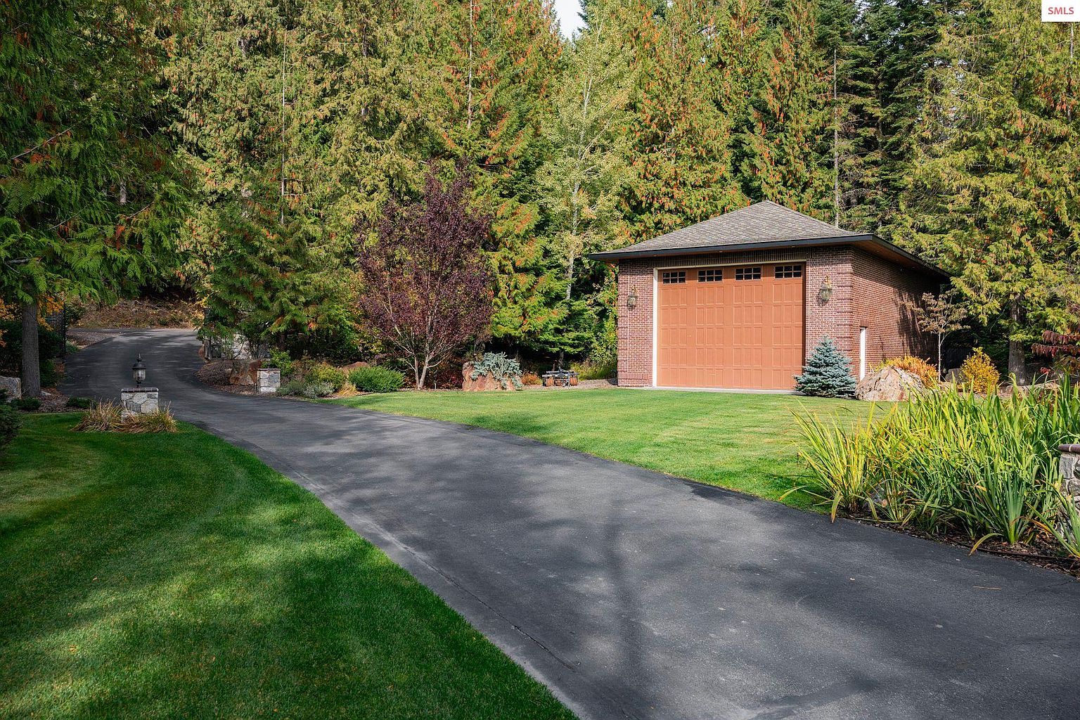 The image showcases a detached brick garage with a brown garage door, set against a backdrop of lush green trees. A paved driveway leads up to the garage, flanked by well-maintained lawns and landscaping. The scene conveys a sense of privacy and well-kept property.
